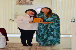 Zwei Frauen auf einer Preisverleihung, die Frau links hält einen Preis, mit einem Rednerpult, einer Wasserflasche, Vorhängen und einem Fotorahmen im Hintergrund.