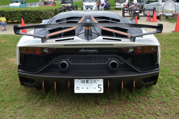 Ein Lamborghini Aventador LP700-4 Superveloce auf dem Goodwood Festival of Speed, mit einem Auto im Vordergrund umgeben von Gras und Verkehrskegeln, Menschen zu Fuß auf der Straßne links und Gebäude, Bäume und einen klaren blauen Himmel im Hintergrund.