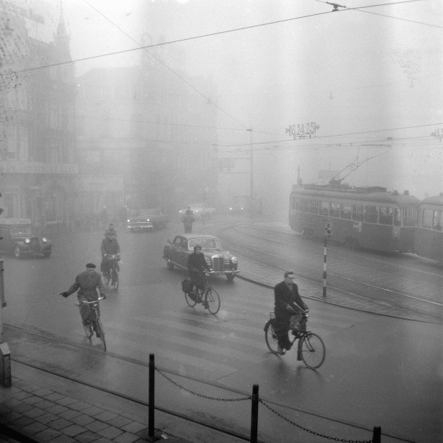 Schwarzes und weißes Foto von Menschen, die Fahrräder auf einer Stadtstraße mit Fahrzeugen, Kettenpfählen und Gebäuden im Hintergrund fahren.