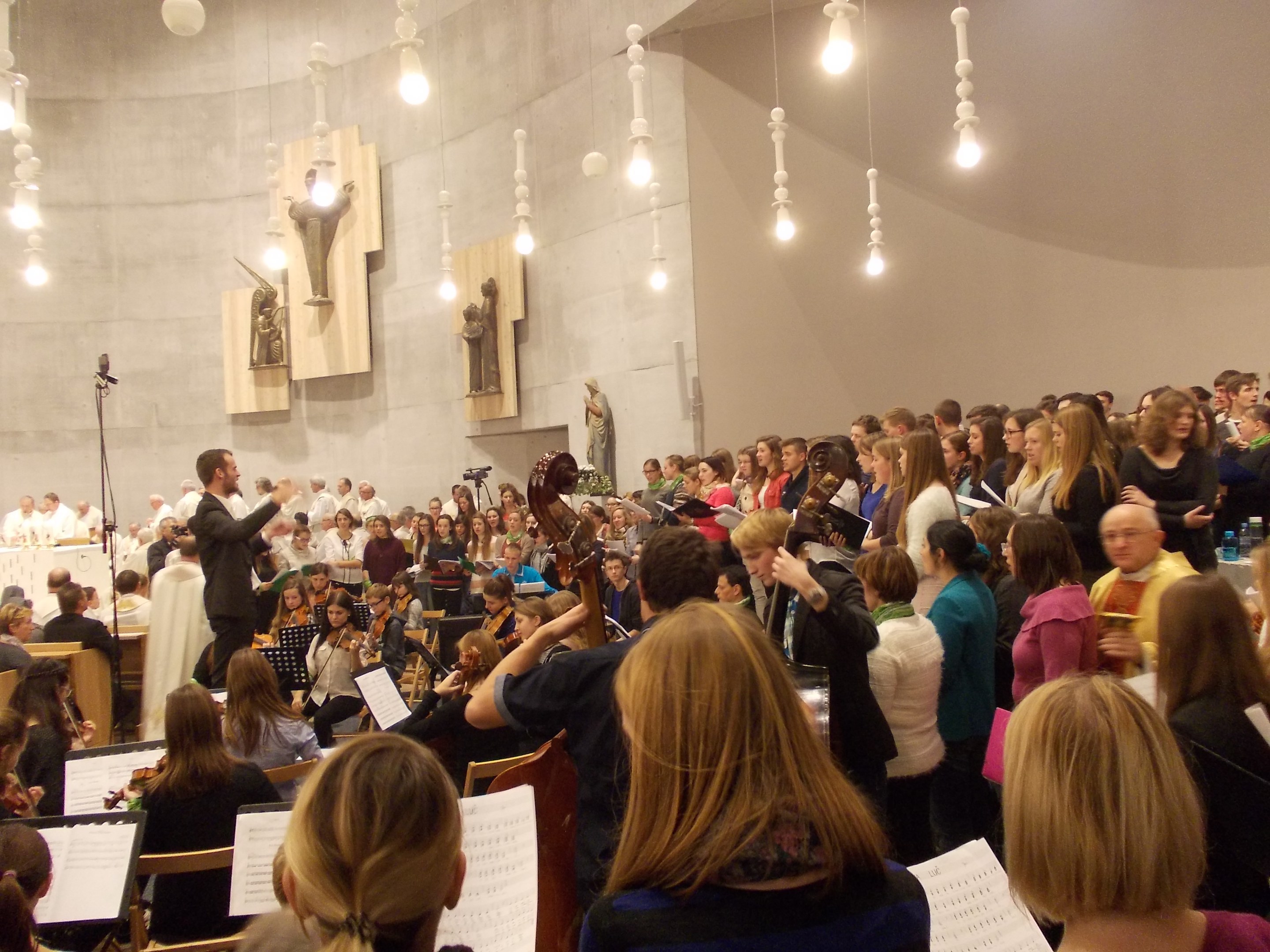 Eine große Gruppe von Menschen steht vor einem Chor, einige halten Musikinstrumente, es gibt Bücher auf Ständern, ein Mikrofon, Statuen an der Wand und Deckenleuchten.