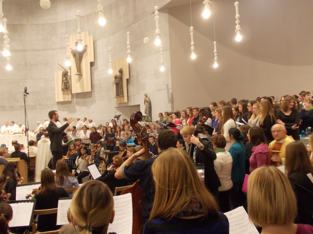 Eine große Gruppe von Menschen steht vor einem Chor, einige halten Musikinstrumente, es gibt Bücher auf Ständern, ein Mikrofon, Statuen an der Wand und Deckenleuchten.