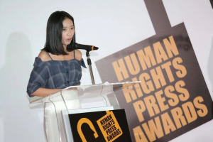 Eine Frau in einem schwarzen Kleid steht an einem Podium mit einem Banner "Human Rights Press Awards" hinter ihr und spricht in ein Mikrofon mit einem ernsten und konzentrierten Gesichtsausdruck.