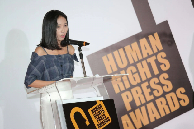 Eine Frau in einem schwarzen Kleid steht an einem Podium mit einem Banner "Human Rights Press Awards" hinter ihr und spricht in ein Mikrofon mit einem ernsten und konzentrierten Gesichtsausdruck.