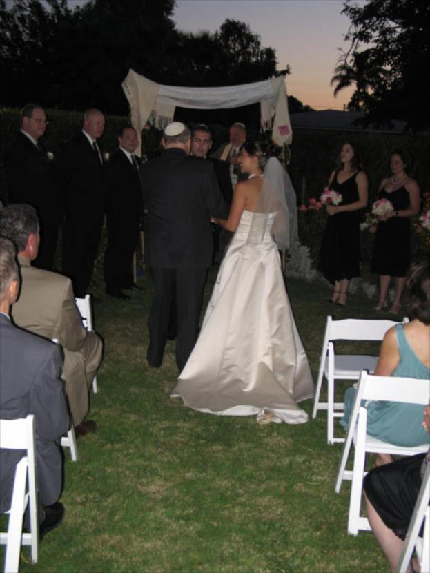 Eine Gruppe von Menschen bei einer Hochzeit, wobei die Frauen rechts Blumensträuße halten und andere auf Stühlen sitzen, während im Hintergrund Bäume zu sehen sind.
