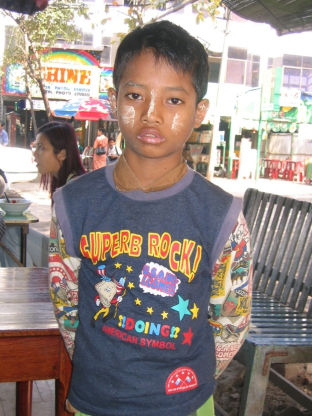 Eine Gruppe von Menschen außerhalb der Stadt, mit einem Jungen in der Mitte, einer Frau auf einem Stuhl links und einem Tisch mit einer Schüssel und einem Löffel davor, mit Geschäften, einer Fahne, einem Baum, Gebäuden und einem Pfahl im Hintergrund.