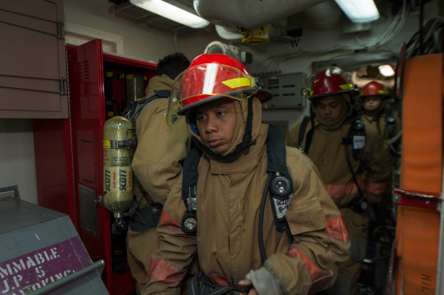 Eine Gruppe von Feuerwehrleuten in Uniformen und Helmen, einer hält einen Feuerlöscher, mit einem metallischen Objekt links und Lichtern, Rohren und anderen Gegenständen im Hintergrund.