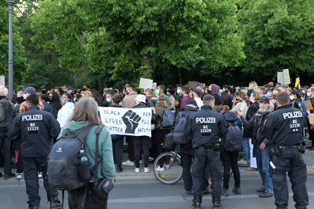 Eine große Gruppe von Menschen nimmt an einem Black Lives Matter-Protest auf der Straße teil, einige halten Schilder und andere tragen Kappen und Taschen, während im Vordergrund ein Fahrrad und im Hintergrund Bäume und ein Pfahl zu sehen sind.