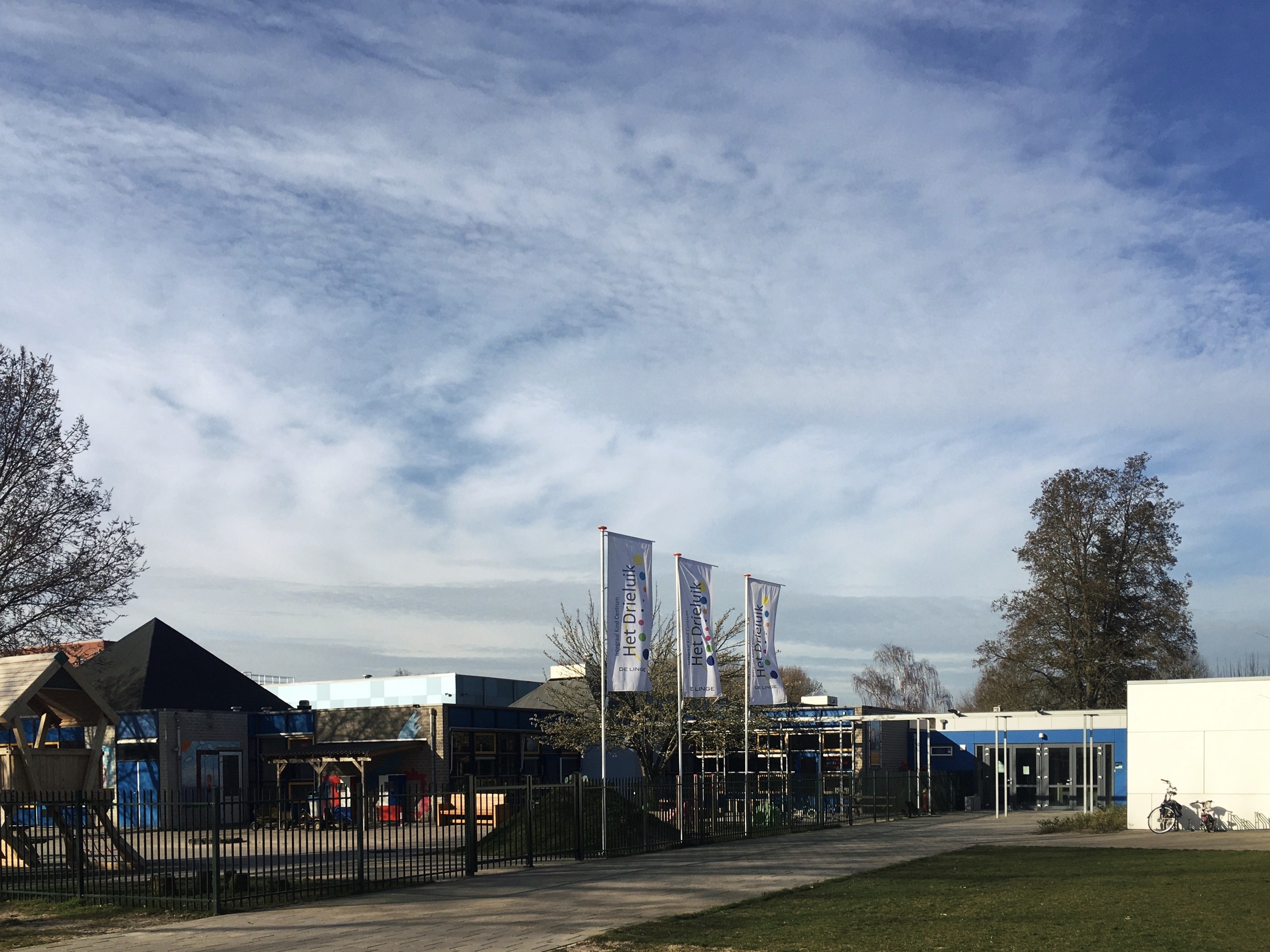 Großes Gebäude mit Metallzaun, Rasenfläche, Weg, Bäumen, Pfosten, Bannern, einem geparkten Fahrrad, nahen Häusern und einem bewölkten Himmel, identifiziert als der neue Spielplatz an der Schule.