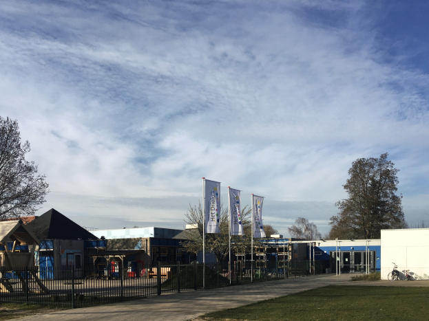 Großes Gebäude mit Metallzaun, Rasenfläche, Weg, Bäumen, Pfosten, Bannern, einem geparkten Fahrrad, nahen Häusern und einem bewölkten Himmel, identifiziert als der neue Spielplatz an der Schule.