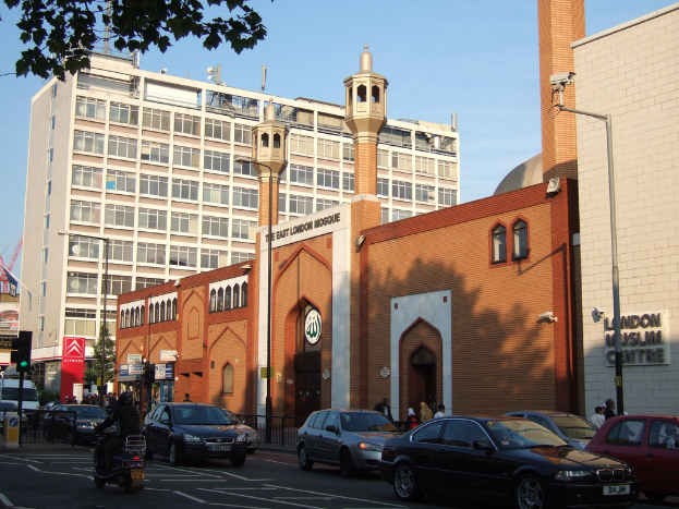 Eine Moschee mit einem Namenbrett an der Wand, Straßenlaternen, eine Ampel, Fahrzeuge auf der Straße, Gruppen von Menschen, die stehen, ein Baum und eine an einem Pfahl angebrachte Überwachungskamera.