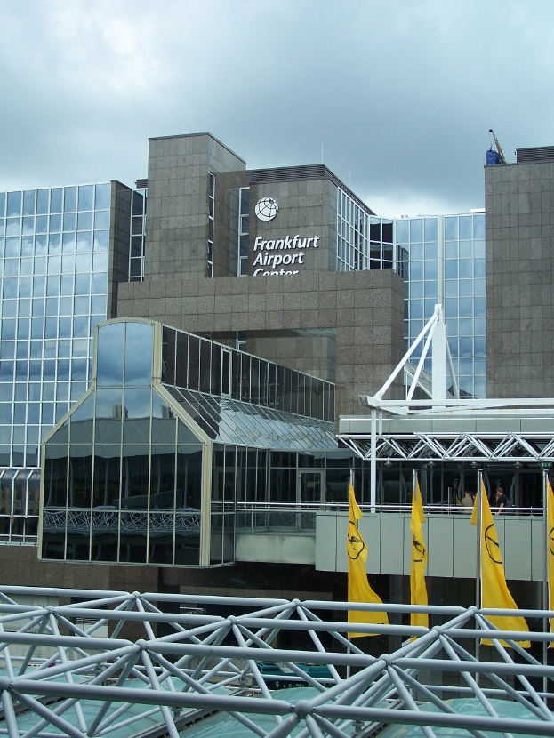 Frankfurter Flughafen in Frankfurt, Deutschland, großes Gebäude mit Glaswänden und Text, umgeben von gelben Flaggen und Eisenstangen, mit einem Himmel voller weißer, flauschiger Wolken im Hintergrund.