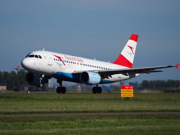Austrian Airlines Airbus A320-200 beim Start am Frankfurt Airport, mit einem Schild im Vordergrund, Gras, Bäumen, Gebäuden und einem klaren blauen Himmel im Hintergrund.
