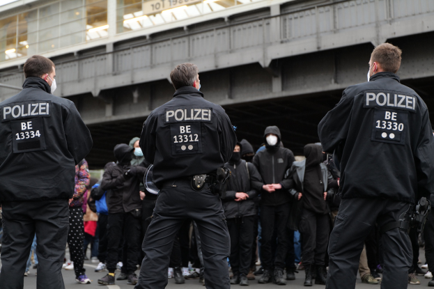 Polizeibeamte in schwarzen Uniformen und Masken vor einer Menge stehend, mit einer Brücke und einem Gebäude im Hintergrund.
