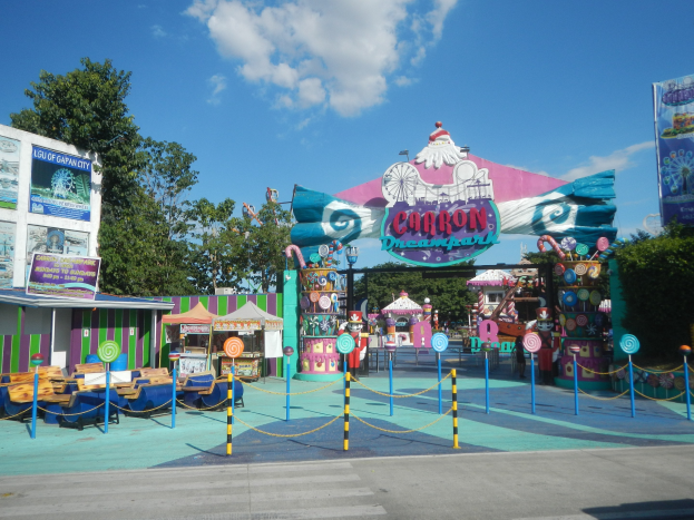 Kirmesfahrgeschäft am Parkeingang mit Straße, Umzäunung, Buden, Bannern, Bäumen und bewölktem Himmel im Hintergrund.