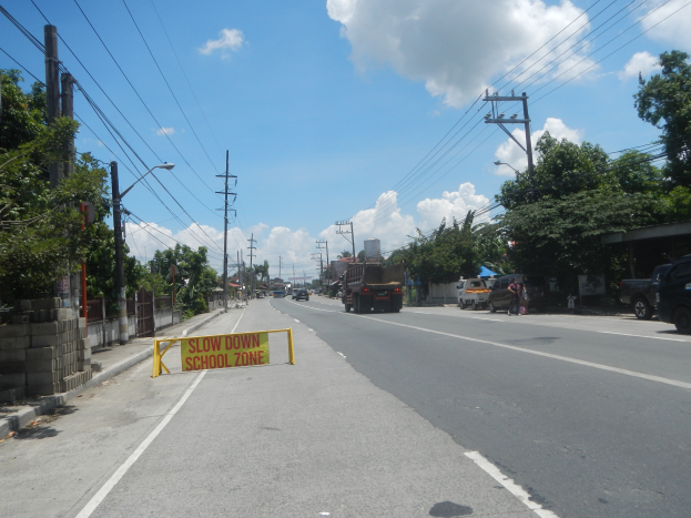 Eine Straße mit Fahrzeugen, ein "Slow Down School Zone"-Schild, Strommasten mit Drähten, Bäume, Laternenpfähle und ein bewölkter Himmel.