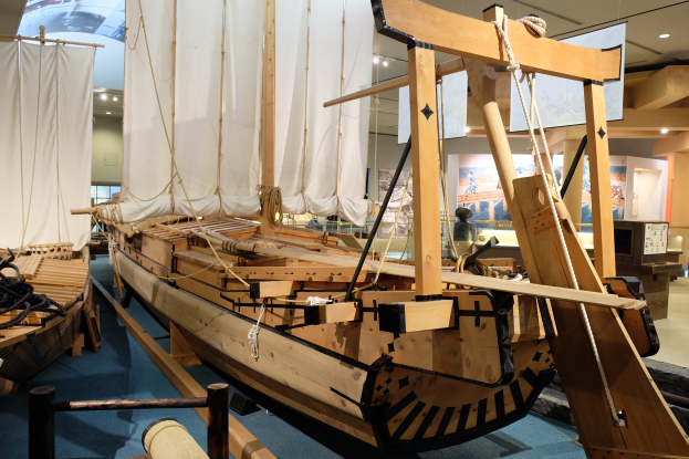 Ein hölzernes Boot, das in einem Museum ausgestellt ist und von Seilen, Pfählen und anderen Gegenständen umgeben ist, mit Brettern, Lampen und einer Wand im Hintergrund.