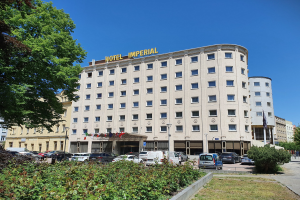 Das Kaiserhotel in Berlin, ein großes Gebäude mit Fenstern, umgeben von Grün, geparkten Autos, Straßenmasten mit Flaggen, unter einem bewölkten Himmel.