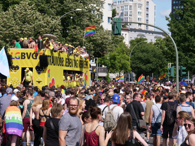 Eine große Menge marschiert auf einer Berliner Straße während eines Umzugs, mit einem gelben Lastwagen im Hintergrund, viele tragen Mützen und Schutzbrillen, einige halten Fahnen, bei bewölktem Himmel mit städtischer Infrastruktur und Gebäuden.