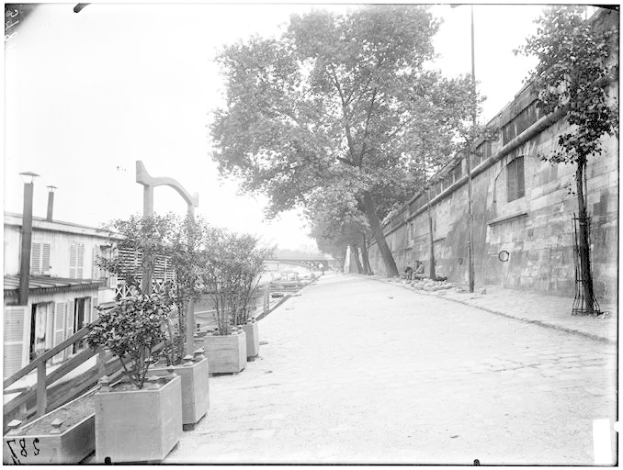 Ein Schwarz-Weiß-Foto einer Straße mit einer Wand auf der rechten Seite, Bäumen und Pflanzen auf der linken Seite und einer Brücke im Hintergrund, mit Text unten, der lautet "New York City, New York, New Jersey, NY, USA, USA - Straßenansicht des New Yorker Staatsgefängnisses".