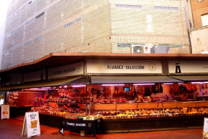 Ein belebter Markt mit verschiedenen Lebensmitteln, beleuchtet von Lichtern und umgeben von Texttafeln, vor Buildings unter einem klaren blauen Himmel.