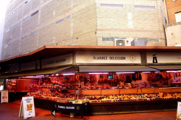 Ein belebter Markt mit verschiedenen Lebensmitteln, beleuchtet von Lichtern und umgeben von Texttafeln, vor Buildings unter einem klaren blauen Himmel.