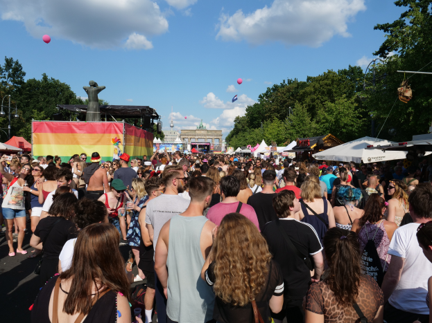 Eine große Menschenmenge, die eine Straße mit Zelten, Bäumen, Pfählen, Laternen und einer Statue entlanggeht, mit Gebäuden im Hintergrund und einem bewölkten Himmel mit Ballons, bei der Christopher Street Day Parade in Berlin.