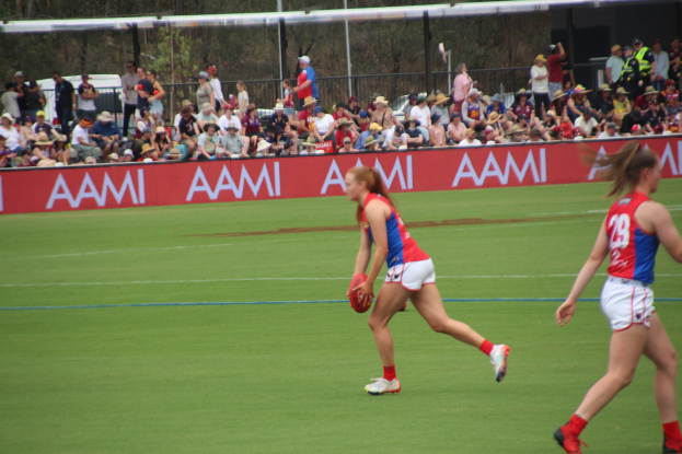 Zwei Frauen spielen Australian Football League auf einem Feld, eine hält einen Ball, mit Zuschauern, Tafeln, Zäunen, Pfählen und Bäumen im Hintergrund.