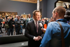 Ein Mann im Anzug und mit Krawatte spricht mit einem Reporter und hält ein Mikrofon in der Hand, während im Hintergrund eine Menschenmenge steht, einige halten Kameras, ein Sofa ist im Vordergrund zu sehen und eine Wand mit einem Rahmen und Lampen dahinter.