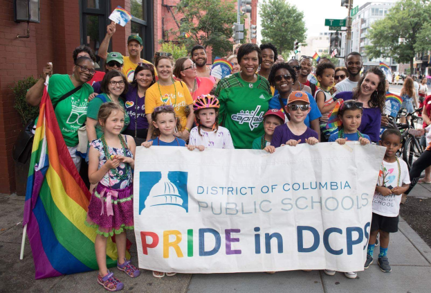 Gruppe von Menschen, die auf der Straße stehen und ein Banner mit der Aufschrift 'District of Columbia Public Schools Pride in DCPS' halten, einige halten eine Fahne, ein Fahrradfahrer, Bäume, Gebäude, Straßenschilder, Ampeln und ein bewölkter Himmel.