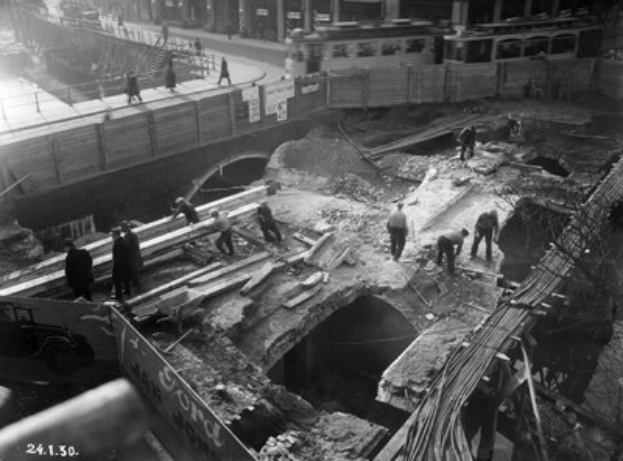 Schwarzes und weißes Foto von Bauarbeitern bei der Reparatur einer Brücke, mit Menschen auf der Brücke und Fahrzeugen auf der Straße im Hintergrund.