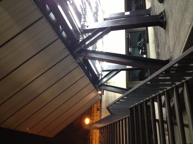 Treppen mit Geländern in der rechten unteren Ecke, Säulen auf der rechten Seite des Bodens, Autos in Gebäuden hinter den Säulen und Lampen auf der linken Seite mit sichtbarer Decke.