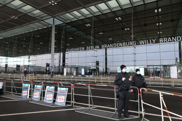 Zwei Polizisten in schwarzen Uniformen stehen vor einem großen Glasgebäude mit Text darauf, mit Absperrungen und Schildern im Vordergrund und anderen Menschen und Gegenständen im Hintergrund.