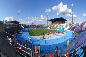Großes Stadion mit Zuschauern bei einem Leichtathletikwettkampf, umgeben von einem Zaun, Laternenmasten, Hütten, Bäumen und Gebäuden unter einem bewölkten Himmel.