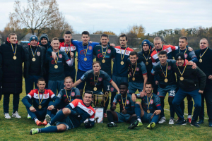 Eine Gruppe von Männern in Medaillen steht auf einem Fußballfeld und hält einen Pokal, umgeben von Bäumen unter einem klaren blauen Himmel.