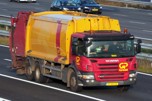 Ein gelber und roter Müllwagen fährt auf einer Autobahn, umgeben von anderen Autos, mit einer Begrenzung und Gras auf beiden Seiten der Straße.