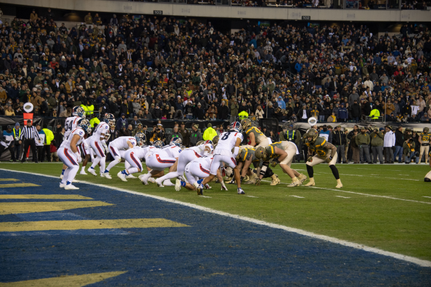 Ein Fußballspiel im Gange mit Spielern in Sportkleidung und Helmen auf dem Feld, Zuschauern, Geländern und Tafeln mit Text im Hintergrund.