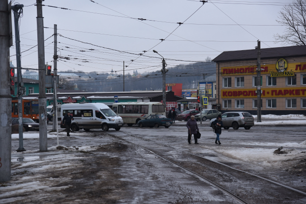 Gruppe von Menschen, die neben einer Bahnschiene auf einer Straße mit Fahrzeugen, Strommästen, Ampeln, Gebäuden, Bäumen und einem Lieferwagen gehen.