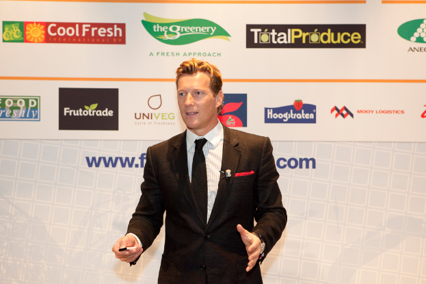 Ein Mann in einer schwarzen Jacke steht und hält ein Objekt, während er spricht, mit einem weißen Banner und einer weißen Tafel mit organisationalen Logos im Hintergrund.