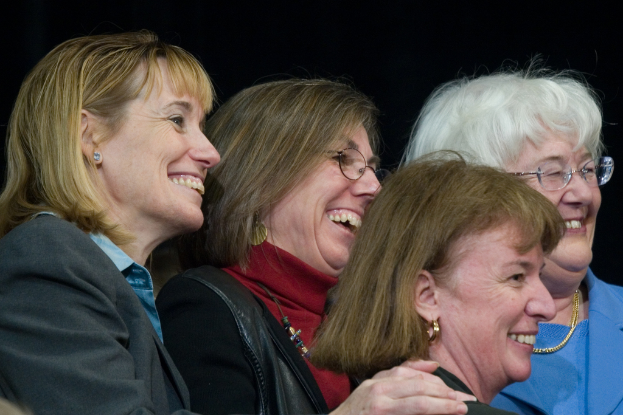 Mehrere ältere Frauen stehen im Vordergrund, löchelnd und nach rechts schauend, mit einem schwarzen Hintergrund dahinter.