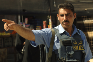 Polizeibeamter in Uniform mit Pistole vor einem Geschäft stehend, mit unscharfen Passanten und Gegenständen im Hintergrund.