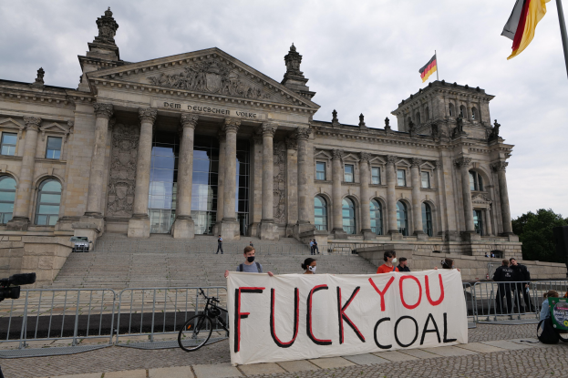 Protestierende halten ein "Fuck You Coal"-Schild vor dem Reichstag in Berlin, mit Bäumen, Fahnenmast und bewölktem Himmel im Hintergrund.
