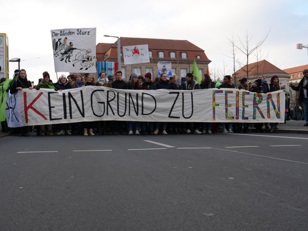 Eine Gruppe von Menschen mit einem Transparent "Kein Grund zu Feiern" protestiert gegen die Sparpolitik der deutschen Regierung auf einer Straße mit Gebäuden, Laternenmasten, Bäumen und einem klaren blauen Himmel im Hintergrund.