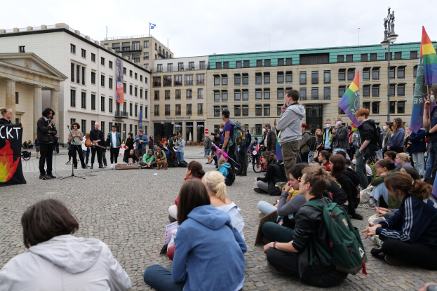 Eine Gruppe von Menschen, die auf dem Boden vor einer Menge mit Fahnen und Spruchbändern sitzt, mit einer Person, die in ein Mikrofon spricht, einer Statue und Gebäuden im Hintergrund während einer anti-schwulen Demonstration in Berlin.