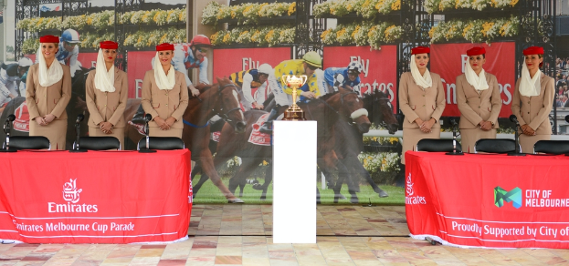 Sechs Flugbegleiterinnen in braunen Anzügen stehen lachend vor der Kamera, mit einem Emirates-Tisch davor und einem Polo-Plakat dahinter, das eine goldene Trophäe auf einem weißen Tisch zeigt.