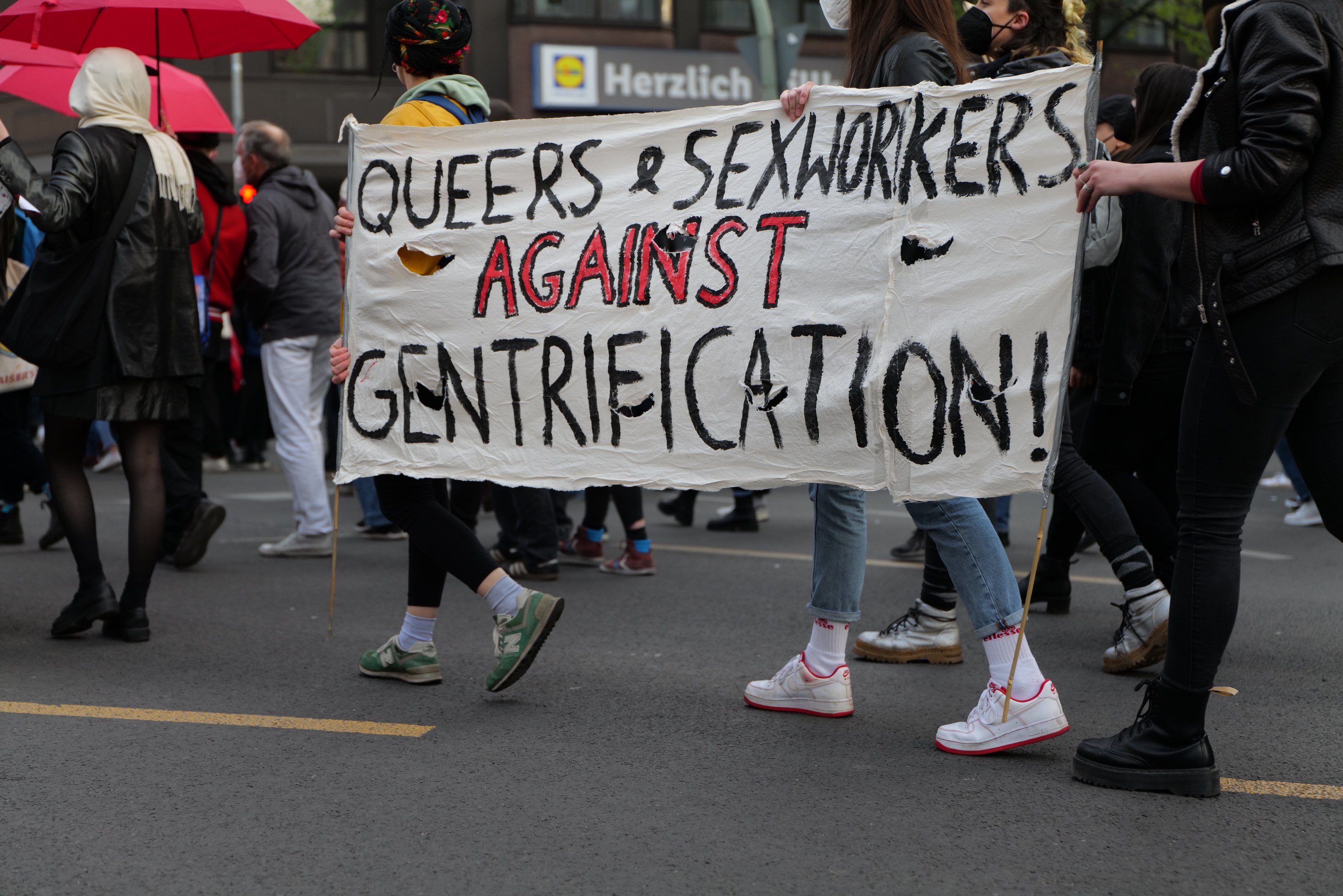 Eine Gruppe von Menschen marschiert auf einer Straße, hält ein Banner mit der Aufschrift "Queers und Sexworker gegen Gentrifizierung", einige tragen Masken und einer hält einen Schirm, vor Bäumen und einem Gebäude mit Fenstern.