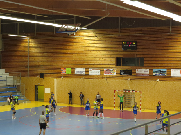Eine Gruppe von Menschen, die Handball in einer Turnhalle mit einem Metallzaun, einem Tor, einer Tür, Texttafeln, einem Anzeigebildschirm, einer Lautsprecherbox, Metallstangen und einem Dach mit Deckenleuchten und Metallrahmen spielen.