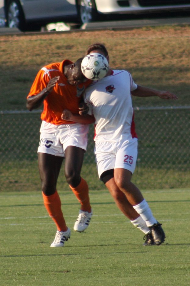 Zwei Spieler auf einem umzäunten Platz, einer im orangen Trikot, der den Ball mit dem Kopf schlägt, mit Autos und Gras im Hintergrund.