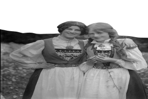 Zwei junge Frauen in traditioneller bayrischer Tracht, mit weißen und schwarzen Kleidern und aufwendiger Stickerei und Perlen, posieren vor einer Kulisse aus üppigen grünen Pflanzen und einem hellen Himmel.