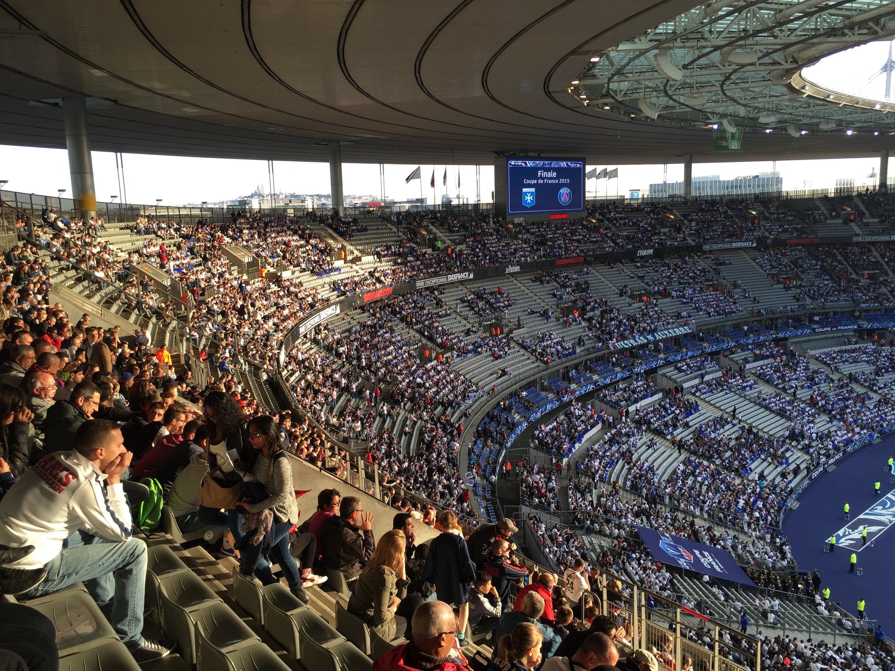 Große Menschenmenge in einem Stadion bei einem Füssballspiel, mit einer Bühne auf der rechten Seite, Fahnen, Masten, einem Bildschirm und der Allianz Arena in München, Deutschland im Hintergrund.