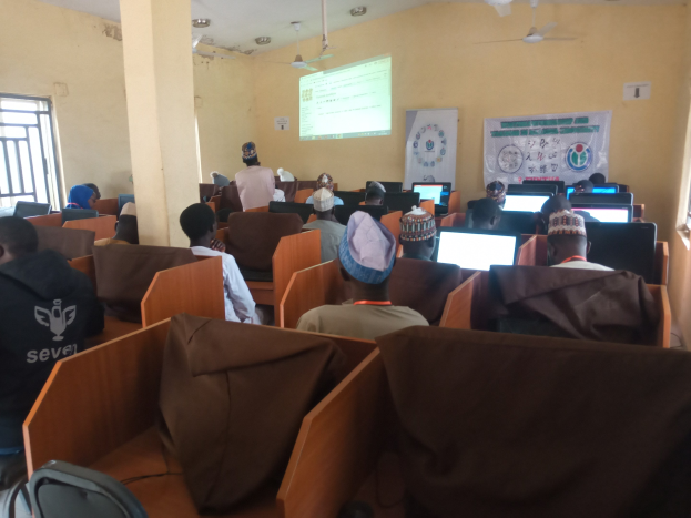 Gruppe von Menschen an Computern in einem Seminarrraum mit Deckenventilatoren, einem Bildschirm, einem Banner und Fenstern an den Seiten.